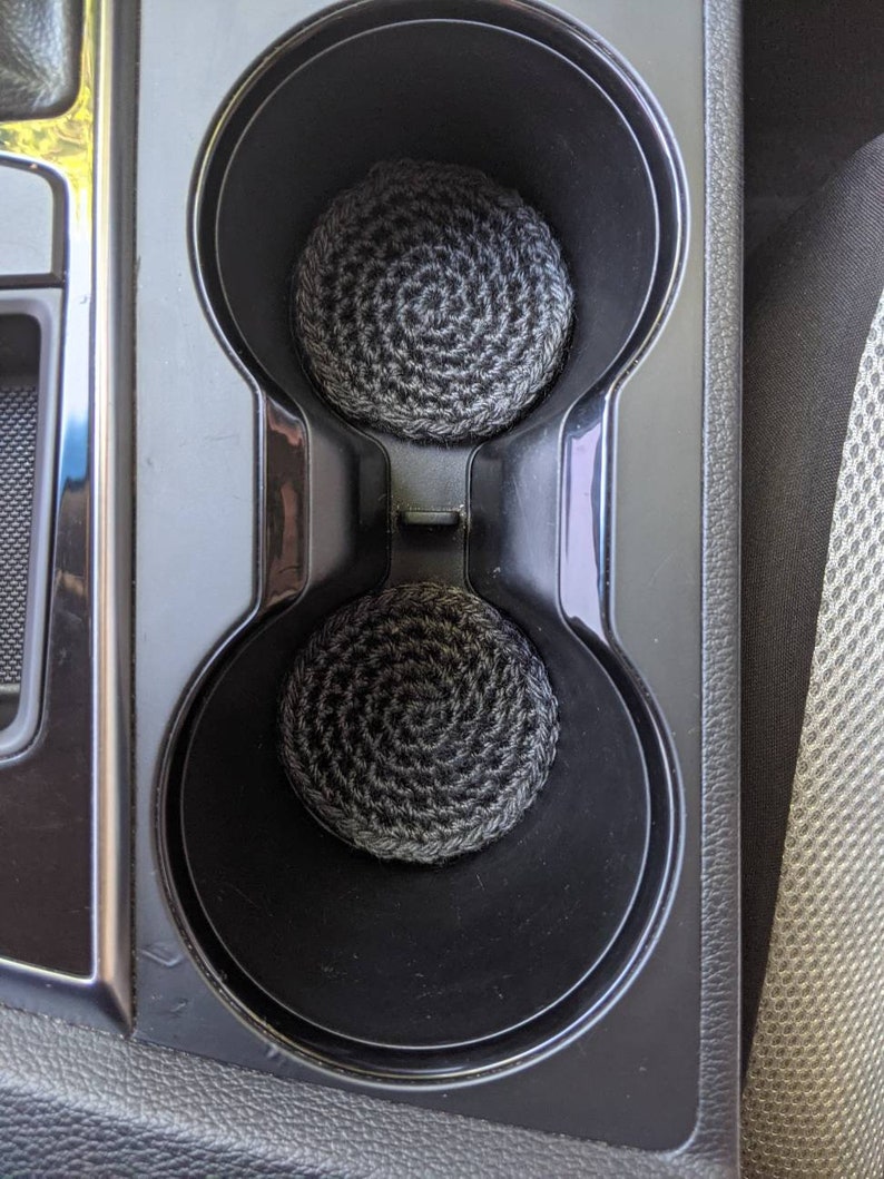 May include: Two black and gray crocheted cup holders in a car's cup holder.