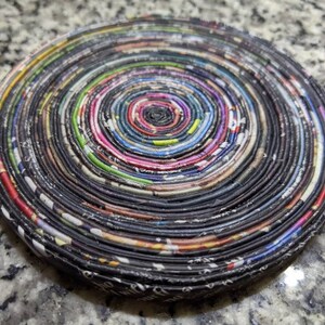 May include: A round, multi-colored coaster made from rolled up newspaper pages. The coaster has a spiral pattern with a dark center and a variety of colors throughout.