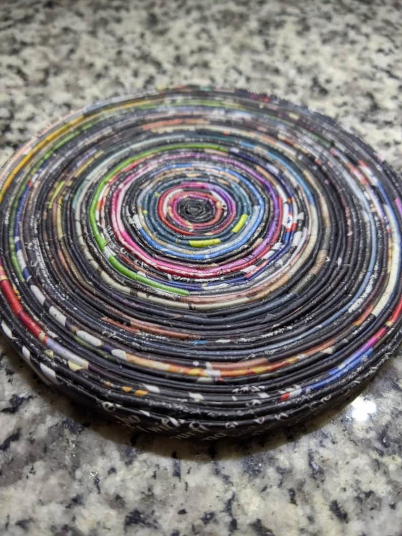 May include: A round, multi-colored coaster made from rolled up newspaper pages. The coaster has a spiral pattern with a dark center and a variety of colors throughout.