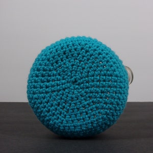 May include: A teal blue crocheted bottle cover. The cover is made of a soft, textured yarn and has a round shape.