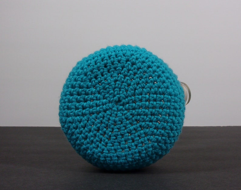 May include: A teal blue crocheted bottle cover. The cover is made of a soft, textured yarn and has a round shape.