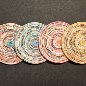 May include: Four round coasters made from recycled paper, each with a different color scheme of concentric circles in shades of pink, blue, yellow, and brown.