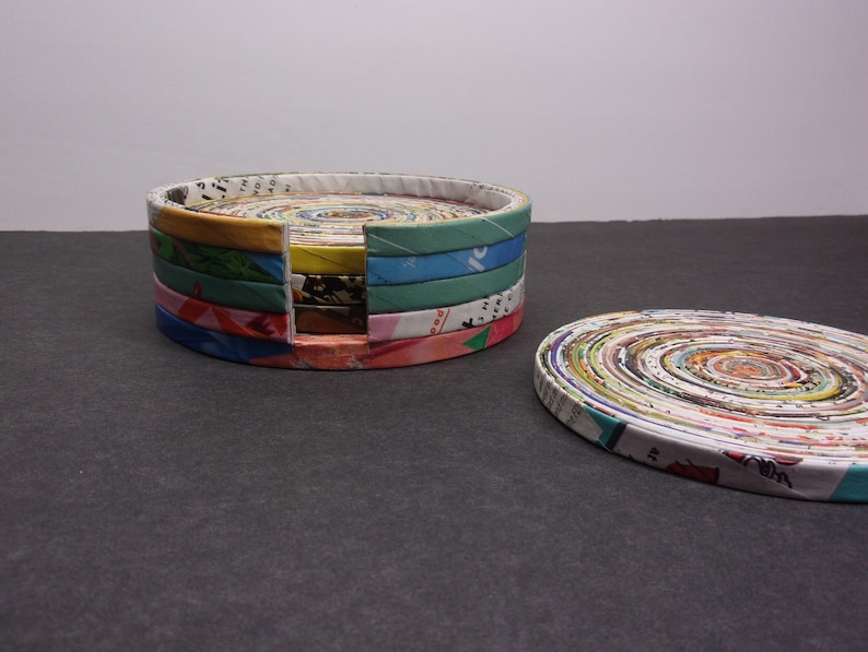 May include: A set of round magazine paper coasters in a variety of colors, including pink, green, yellow, and blue. The coasters are stacked in a tower with a gap between the top and bottom layers. The coasters are made from rolled up magazine pages.