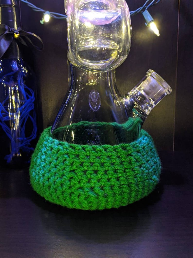 May include: A green crocheted cozy for a glass water pipe with a clear glass bowl and a separate glass mouthpiece.