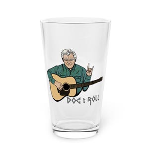 May include: A clear glass pint glass featuring a cartoon illustration of a man playing an acoustic guitar. The man is wearing a green shirt and has grey hair. The text "Doc & Roll" is printed below the image.