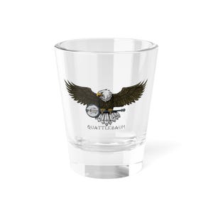 Puede incluir: Un vaso de chupito de vidrio transparente con un gráfico de un águila calva sosteniendo un banjo. El águila está volando con las alas extendidas. El texto "Quattlebaum" está impreso debajo del águila.