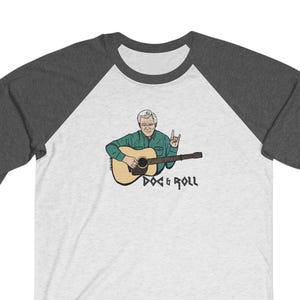 May include: A white and grey baseball raglan t-shirt with a cartoon illustration of a man playing a guitar. The text "Doc & Roll" is printed below the image.