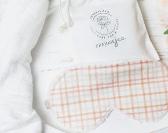 May include: A white cotton sleep mask with a pink and white plaid pattern. The mask is packaged in a white drawstring bag with the text "Frankie & Co. Self Care for a Frankie & Co." printed on the front.