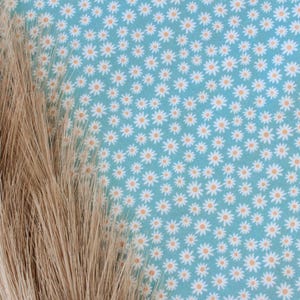 K&ouml;nnte beinhalten: Nahaufnahme eines blauen Stoffes mit einem wei&szlig;en G&auml;nsebl&uuml;mchenmuster. Der Stoff ist strukturiert und hat einen dezenten Glanz. Im Vordergrund ist ein B&uuml;ndel getrockneter Weizenhalme teilweise sichtbar.