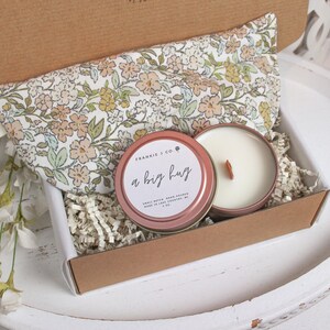 Puede incluir: Una caja de regalo que contiene una almohada para los ojos con estampado floral, una vela en una lata rosa con el texto "a big hug" y papel triturado. La vela tiene una mecha de madera. La caja es marr&oacute;n y blanca.
