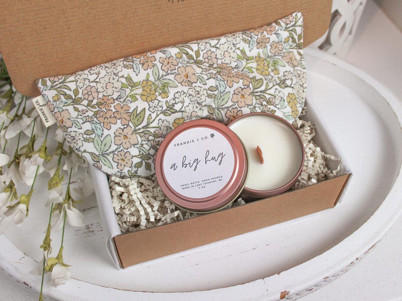 Puede incluir: Una caja de regalo que contiene una almohada para los ojos con estampado floral, una vela en una lata rosa con el texto "a big hug" y papel triturado. La vela tiene una mecha de madera. La caja es marr&oacute;n y blanca.