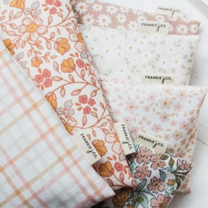 May include: Close-up of several fabric pieces with floral and plaid patterns. The fabrics feature a variety of colors, including peach, pink, and white. Each piece has a tag that reads "FRANKIE jco."