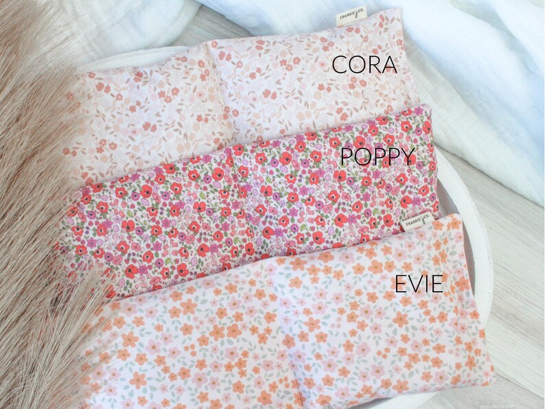 May include: Three floral print rectangular pillows in a white tray. The top pillow is light pink with small flowers, the middle pillow is red and pink with small flowers, and the bottom pillow is white with orange flowers. The pillows have the names "Cora", "Poppy", and "Evie" printed on them.