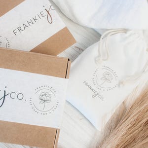 K&ouml;nnte beinhalten: Zwei braune Pappkartons mit wei&szlig;en Etiketten. Die Etiketten zeigen ein Blumenmuster und den Text "Frankie jco. Self care for a cause". Im Hintergrund befindet sich ein wei&szlig;er Kordelbeutel mit einem &auml;hnlichen Blumenmuster und dem Text "Frankie jco. Self care for a cause".