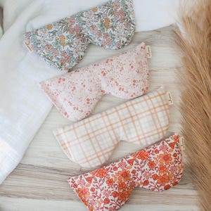 May include: Four eye masks with floral patterns in shades of pink, orange, and blue, and a plaid design. The masks are displayed on a light wooden surface. Each mask is filled with a soft material.