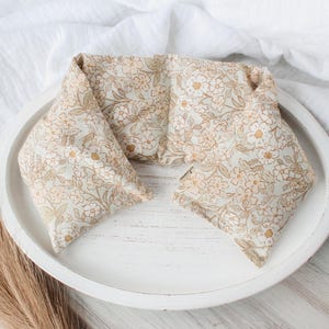 May include: A floral patterned neck wrap in beige and gold tones, resting on a white wooden tray. The wrap is folded into a rectangular shape, with a soft, textured appearance. The background includes a white cloth and dried grass.