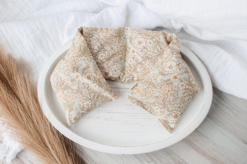 May include: A floral patterned neck wrap in beige and gold tones, resting on a white wooden tray. The wrap is folded into a rectangular shape, with a soft, textured appearance. The background includes a white cloth and dried grass.