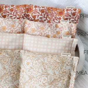 May include: Four rectangular fabric items with floral and gingham patterns. The top item is Phoebe, with orange and red flowers. Primrose has peach and yellow flowers. Fern is a peach gingham. Meadow has white and brown flowers. Each item has a Frankie & Co. tag.