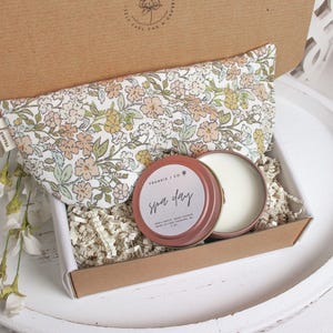 Puede incluir: Caja de regalo abierta con una almohada para los ojos con estampado floral, una peque&ntilde;a lata redonda con la inscripci&oacute;n "Spa Day" y otra lata con una sustancia blanca. La caja es marr&oacute;n y est&aacute; llena de papel triturado.