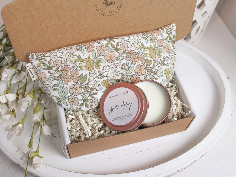 Puede incluir: Caja de regalo abierta con una almohada para los ojos con estampado floral, una peque&ntilde;a lata redonda con la inscripci&oacute;n "Spa Day" y otra lata con una sustancia blanca. La caja es marr&oacute;n y est&aacute; llena de papel triturado.