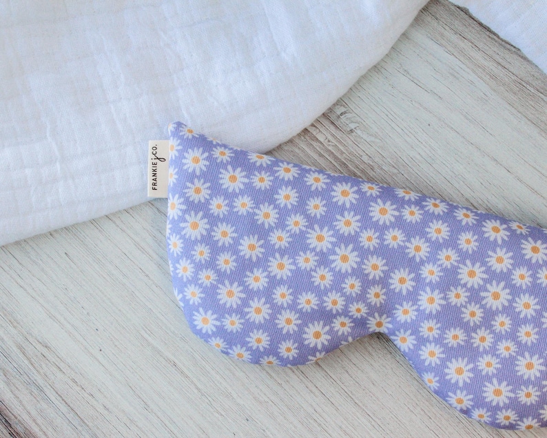 K&ouml;nnte beinhalten: Eine Lavendel-Augenmaske mit einem wei&szlig;en G&auml;nsebl&uuml;mchen-Druck. Die Augenmaske hat ein Etikett mit der Aufschrift "Frankie & Co."