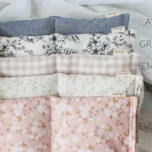 May include: Five fabric items with various patterns and colours are stacked. The top one is a solid blue-grey, followed by a black and white floral print, a beige gingham, a white floral print, and a pink floral print. Each item has a tag that says "frankie & co.".