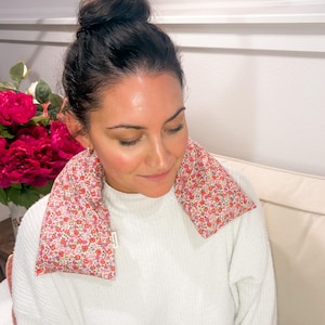 May include: A pink floral print neck wrap rests on a person's shoulders. The wrap is filled with a soft material and is designed to provide comfort. The background includes a white sweater and a bouquet of pink flowers.