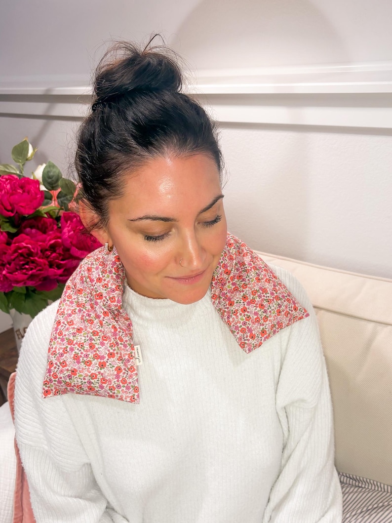 May include: A pink floral print neck wrap rests on a person's shoulders. The wrap is filled with a soft material and is designed to provide comfort. The background includes a white sweater and a bouquet of pink flowers.