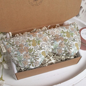 Puede incluir: Una m&aacute;scara para los ojos con estampado floral y una peque&ntilde;a lata se presentan en una caja de cart&oacute;n abierta. La m&aacute;scara para los ojos tiene un fondo blanco con flores melocot&oacute;n, amarillas y verdes. La lata es de color rosa apagado con texto blanco.