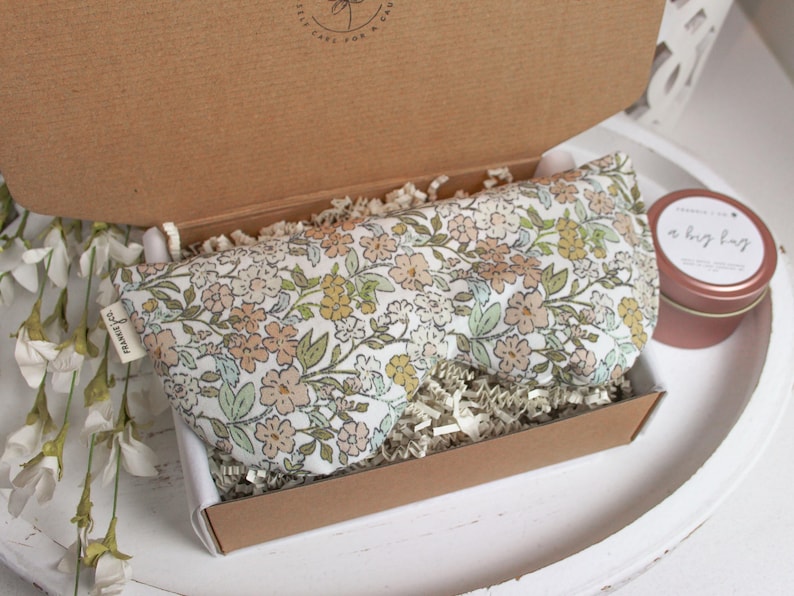 Puede incluir: Una m&aacute;scara para los ojos con estampado floral y una peque&ntilde;a lata se presentan en una caja de cart&oacute;n abierta. La m&aacute;scara para los ojos tiene un fondo blanco con flores melocot&oacute;n, amarillas y verdes. La lata es de color rosa apagado con texto blanco.