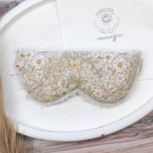 Puede incluir: M&aacute;scara para ojos con estampado floral y un interior blanco y suave. La m&aacute;scara presenta un fondo azul claro con un patr&oacute;n de flores blancas y beige y hojas verdes. Dise&ntilde;ada para bloquear la luz.