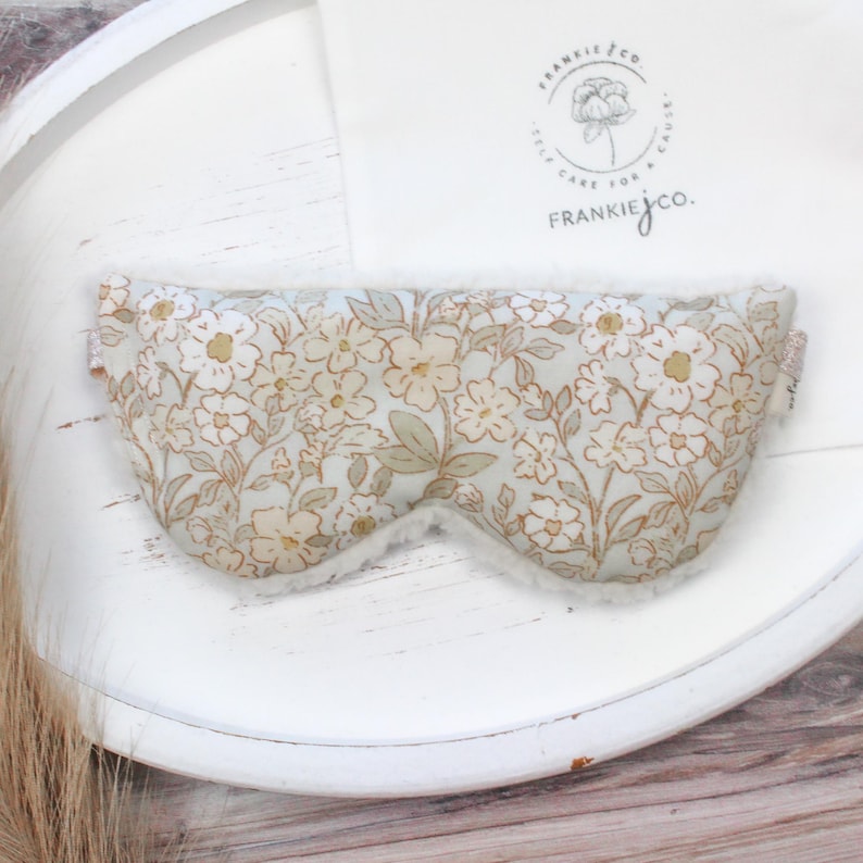 Puede incluir: M&aacute;scara para ojos con estampado floral y un interior blanco y suave. La m&aacute;scara presenta un fondo azul claro con un patr&oacute;n de flores blancas y beige y hojas verdes. Dise&ntilde;ada para bloquear la luz.