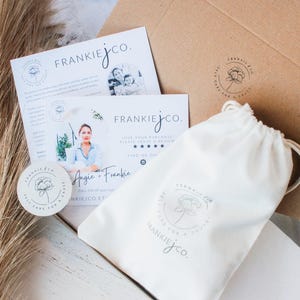 May include: An overhead shot of a product package with branding. The package includes a white drawstring bag, a round sticker, and two cards with the text "FRANKIEJCO." and a floral logo. A brown cardboard box is also visible.