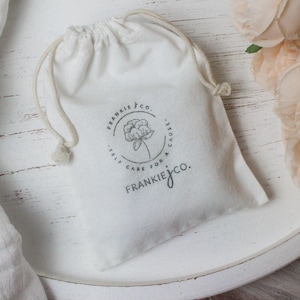 May include: A white drawstring bag with the Frankie & Co. logo, featuring a floral design and the text "Self Care for a Cause." The bag is on a white surface, with a white cloth and peach-colored flowers in the background.
