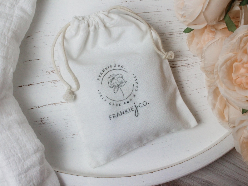 May include: A white drawstring bag with the Frankie & Co. logo, featuring a floral design and the text "Self Care for a Cause." The bag is on a white surface, with a white cloth and peach-colored flowers in the background.