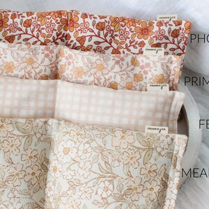 May include: Four fabric-covered pillows with floral and gingham patterns in shades of orange, pink, and beige. The pillows are labeled "Phoebe", "Primrose", "Fern", and "Meadow". Each pillow has a small tag that says "frankie jco."