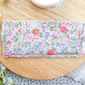 K&ouml;nnte beinhalten: Eine wei&szlig;e Augenmaske mit einem Blumenmuster mit rosa, roten und blauen Blumen. Die Augenmaske hat einen wei&szlig;en, flauschigen Rand.