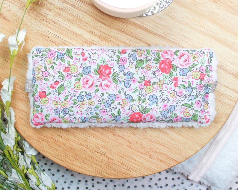 K&ouml;nnte beinhalten: Eine wei&szlig;e Augenmaske mit einem Blumenmuster mit rosa, roten und blauen Blumen. Die Augenmaske hat einen wei&szlig;en, flauschigen Rand.
