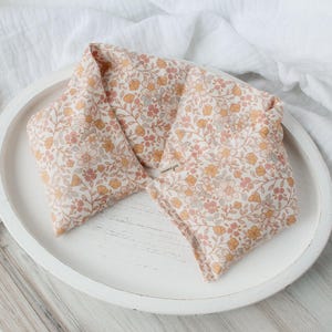 May include: A floral patterned heat pack in shades of peach, yellow, and gray, resting on a white, oval-shaped tray. The fabric features a delicate botanical design. The tray sits on a white surface, with a dried plant stem in the background.