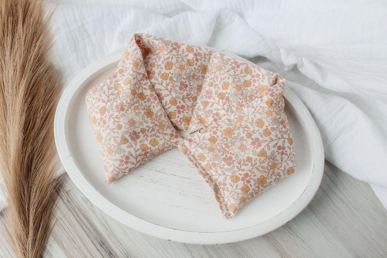 May include: A floral patterned heat pack in shades of peach, yellow, and gray, resting on a white, oval-shaped tray. The fabric features a delicate botanical design. The tray sits on a white surface, with a dried plant stem in the background.