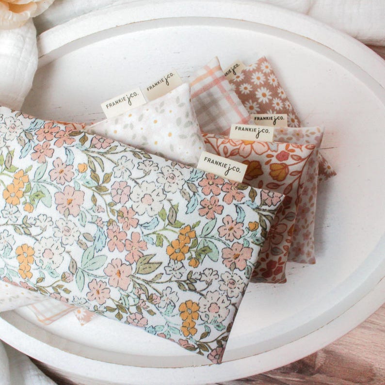 May include: A collection of floral print fabric pouches in various patterns and colors, including pink, orange, and white, arranged on a white tray. Each pouch has a small tag that reads "FRANKIEJ.CO."