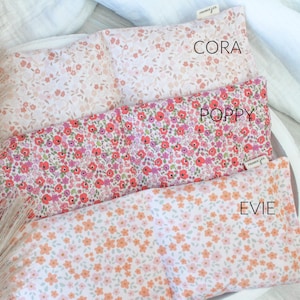 May include: Three rectangular pillows with floral patterns, displayed in a white tray. The top pillow is pale pink with tiny flowers, the middle one is red and pink, and the bottom one is white with orange flowers. The pillows are labelled "Cora", "Poppy", and "Evie".