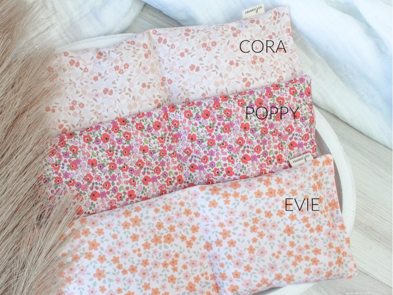 May include: Three rectangular pillows with floral patterns, displayed in a white tray. The top pillow is pale pink with tiny flowers, the middle one is red and pink, and the bottom one is white with orange flowers. The pillows are labelled "Cora", "Poppy", and "Evie".