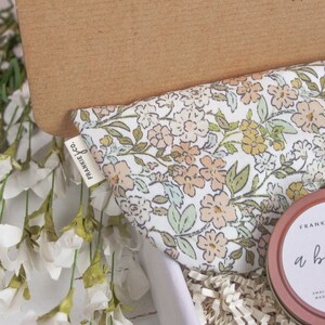 Puede incluir: Una caja de regalo con tela estampada floral, una lata redonda con el texto "a big hug" y una etiqueta que dice "frankie j co.". La caja est&aacute; llena de papel blanco y rodeada de flores blancas.