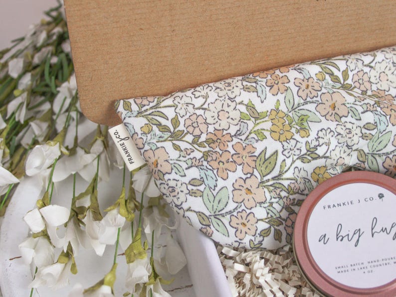 Puede incluir: Una caja de regalo con tela estampada floral, una lata redonda con el texto "a big hug" y una etiqueta que dice "frankie j co.". La caja est&aacute; llena de papel blanco y rodeada de flores blancas.