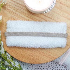 K&ouml;nnte beinhalten: Wei&szlig;er, flauschiger Schlafmaske mit einem silbernen Glitzere&lambda;&alpha;&sigma;&tau;&iota;&kappa;ό. Die Schlafmaske liegt auf einer Holzoberfl&auml;che.