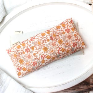May include: A rectangular floral print eye pillow in shades of peach, yellow, and gray, resting on a white tray. The pillow features a delicate pattern of small flowers and leaves. The tray is surrounded by white fabric and peach-colored roses.