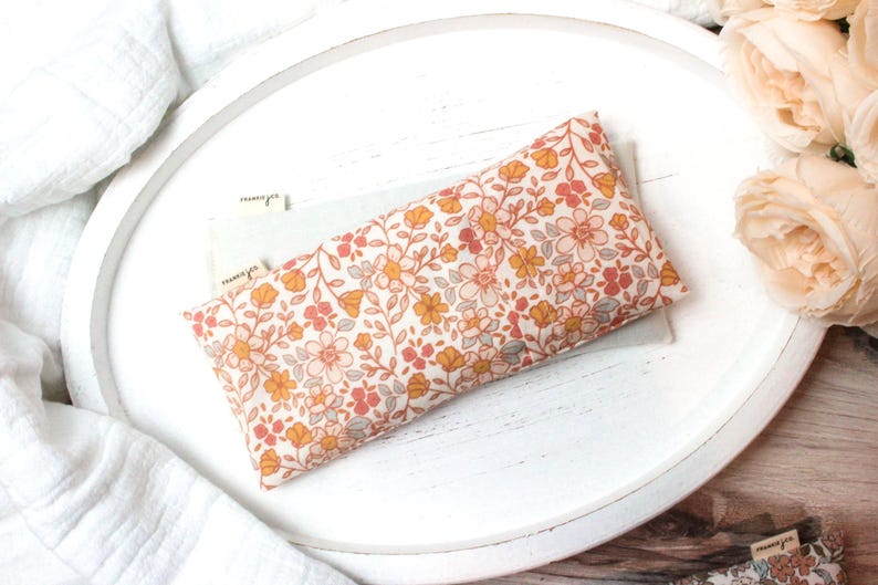 May include: A rectangular floral print eye pillow in shades of peach, yellow, and gray, resting on a white tray. The pillow features a delicate pattern of small flowers and leaves. The tray is surrounded by white fabric and peach-colored roses.