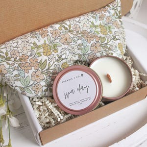 Puede incluir: Un set de regalo para un d&iacute;a de spa en una caja de cart&oacute;n. El set incluye una almohada para los ojos con estampado floral, una vela en una lata de oro rosa con las palabras "Spa Day" y papel triturado. La vela tiene una mecha de madera.