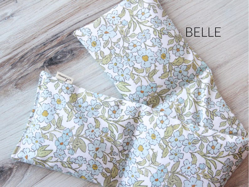 May include: Two rectangular fabric items with a floral pattern. The pattern features light blue flowers, green leaves, and yellow centers on a white background. The word "BELLE" is printed in black. A small tag reads "frankie & co."